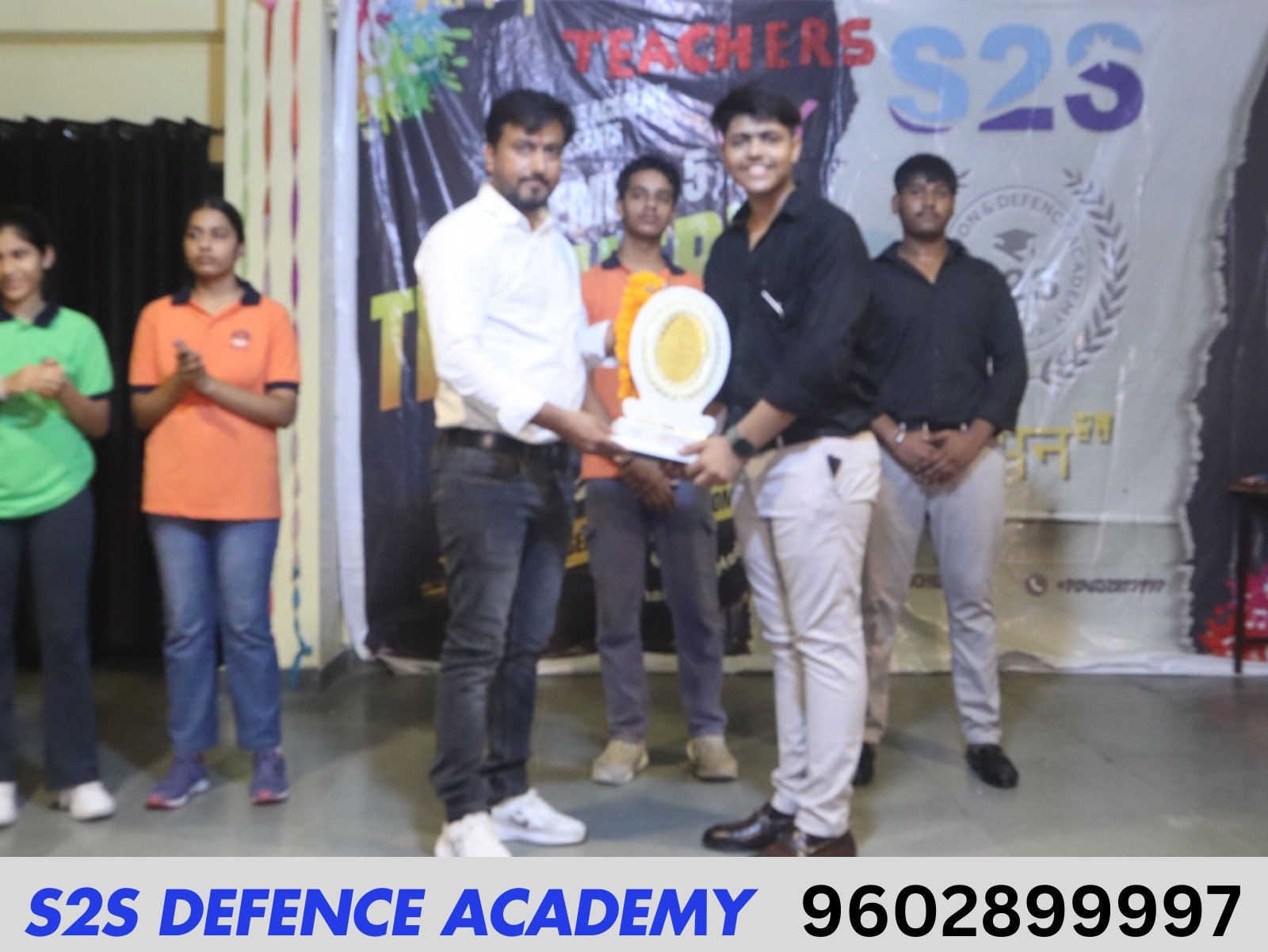 A young man in a black shirt receives an award from another man in a white shirt, surrounded by clapping students in s2s defence academy